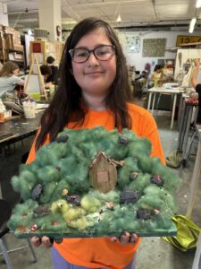 A girl holding up a cake with trees and houses on it.