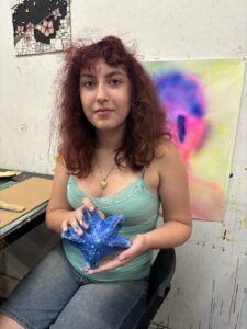 A woman sitting in front of a painting holding something blue.