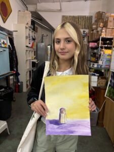 A girl holding up a painting of a lighthouse.