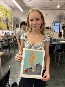 A girl holding up an art project in front of other students.