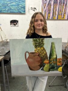 A girl holding up a painting of fruit and a pitcher.
