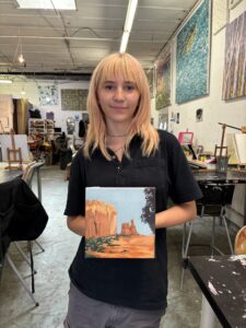 Here's an alt tag for the image: Young artist holding desert landscape painting.
