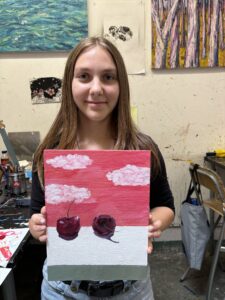 Girl holding painting of cherries and clouds.