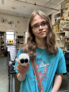 A young person holding a small, round, blue object with big eyes in a cluttered room.