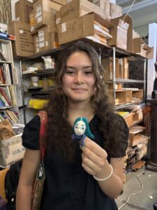 Young woman holding a small handmade doll in a cluttered workspace.