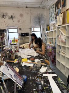 Two women working on art projects in a cluttered studio filled with supplies and artwork.