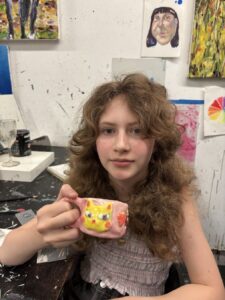 Young woman with curly hair holding a colorful mug in an art studio.