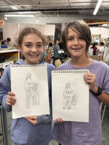 Two children proudly show their pencil sketches in an art class.