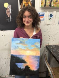 Young woman proudly holds her sunset landscape painting in an art studio.