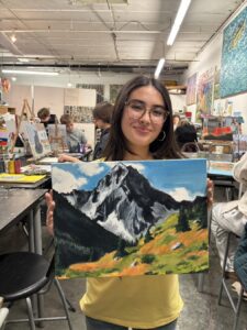 A woman proudly holds her mountain landscape painting in an art studio.