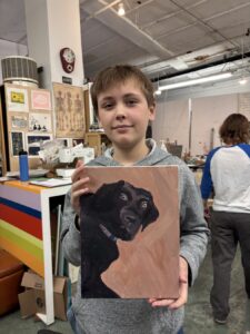A boy proudly holding a painting of a black dog indoors.