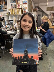Girl proudly displays her painting of a city skyline at sunset.