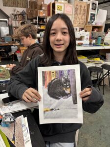 A girl proudly holds a drawing of a black cat in an art classroom.