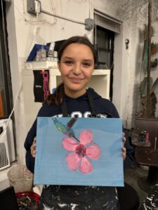 A smiling girl proudly holds a blue paper with a pink flower craft.