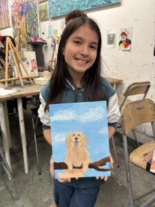 Girl proudly shows her painting of a golden retriever against a blue sky.