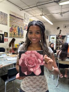 A smiling woman holding a painting of a pink rose in an art studio.