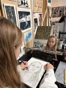 A young girl sketching her own portrait in front of a mirror.