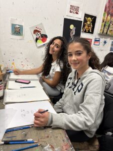 Two girls sitting at desks writing, smiling at the camera.